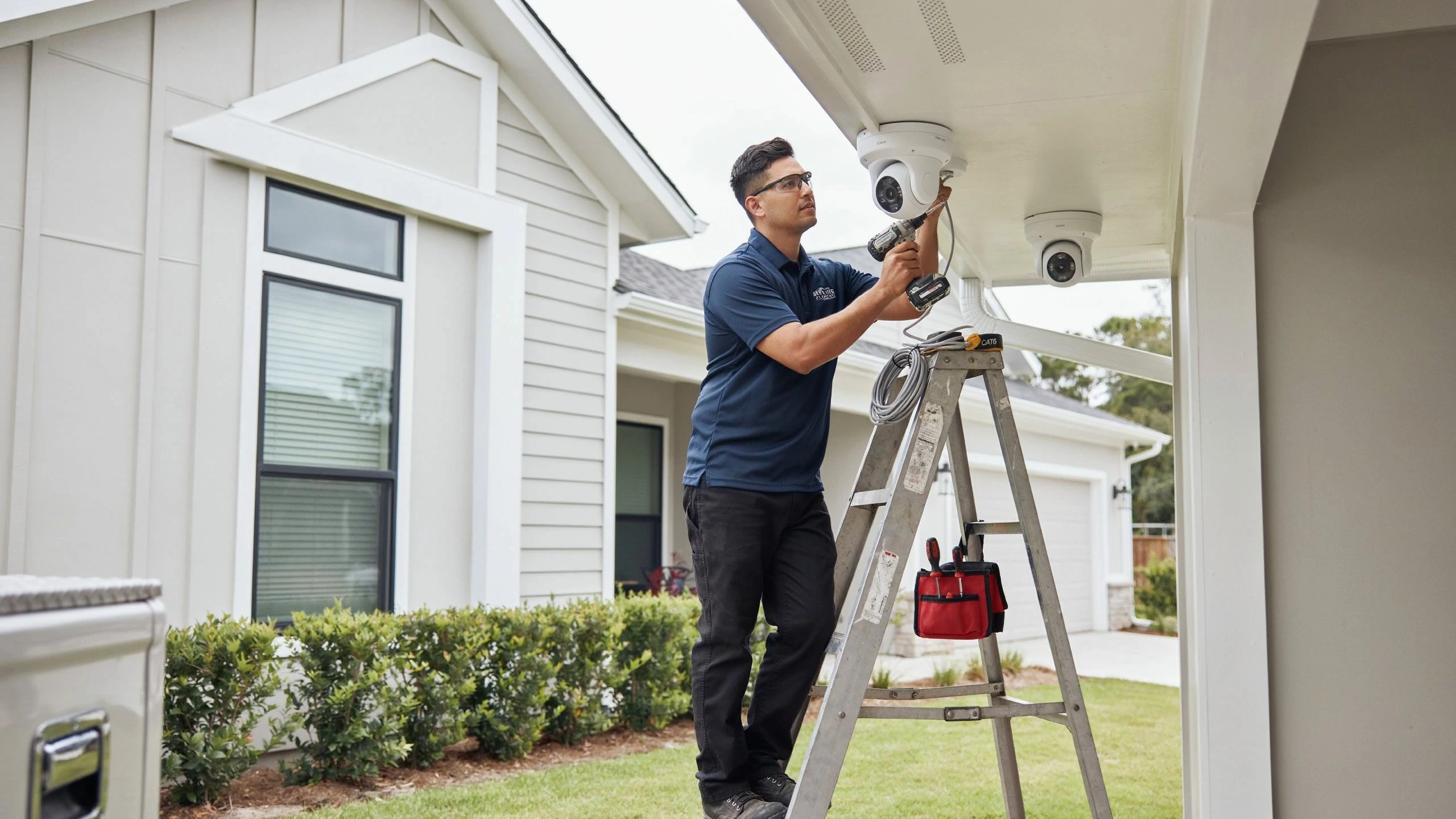 Technician on a ladder installing a dome security camera under a home's covered porch, with a second camera visible nearby.