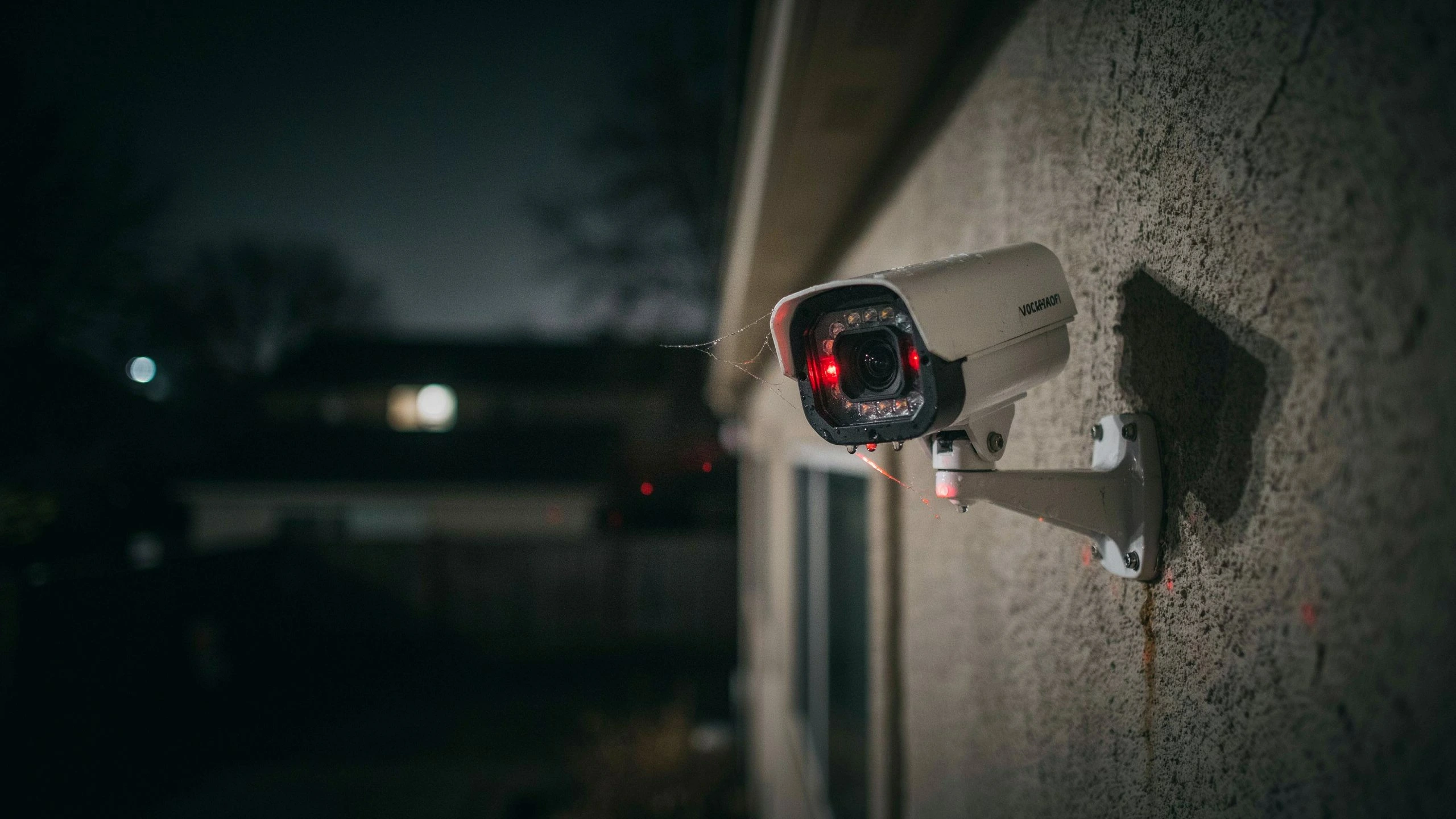 Side view of a security camera at night with glowing red infrared sensors and a cobweb stretching across the lens area.