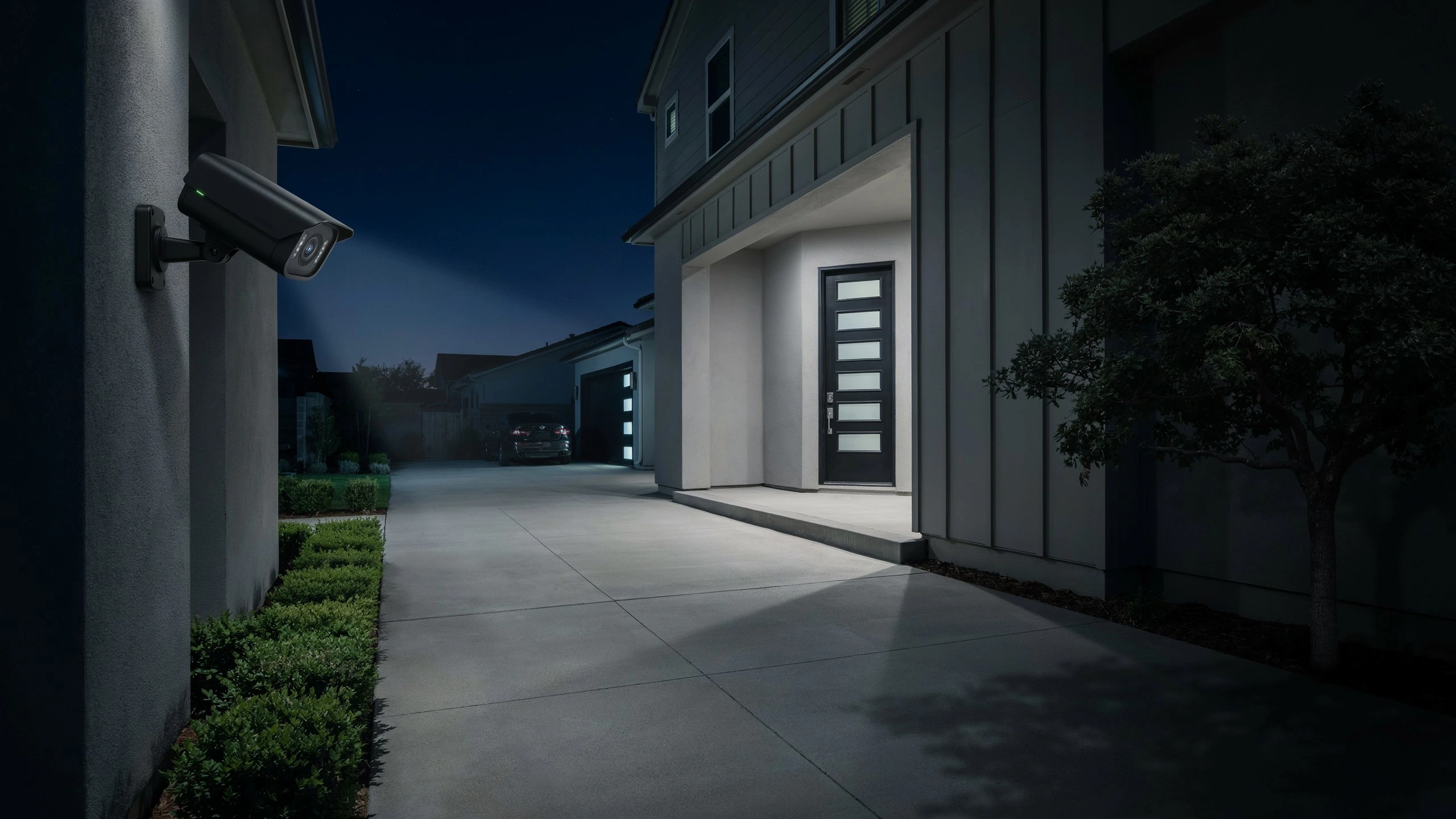 Black bullet security camera mounted on an exterior wall, casting a beam of light down a driveway toward a house at night.